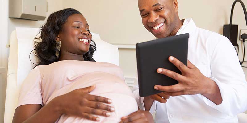 A pregnant woman and her healthcare provider smile at a tablet, which presumbably contains test results or other health information.