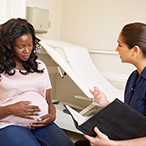 A pregnant African-American woman is with her healthcare provider, who is speaking and holding a notebook.