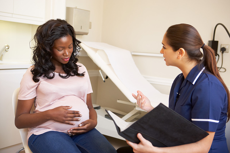 A pregnant African-American woman is with her healthcare provider, who is speaking and holding a notebook.