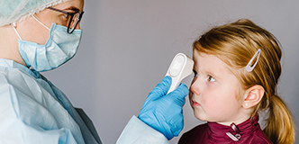 Medical professional wearing mask and gloves scanning a child’s forehead with a digital thermometer.