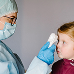 Medical professional wearing mask and gloves scanning a child’s forehead with a digital thermometer.