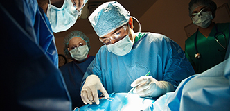 Doctors and nurses in surgical garb standing over a patient’s draped torso.