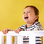 Infant standing behind crib rail, crying.