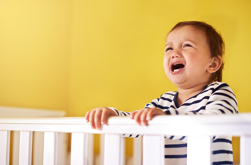 Infant standing behind crib rail, crying.