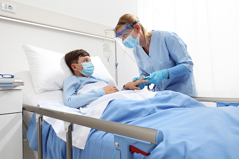 Masked child lying in hospital bed while nurse checks oxygen level on a finger pulse oximeter.