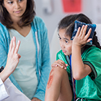 Girl with skinned knee holding ice pack to her head looking at healthcare provider holding up three fingers while woman looks on.