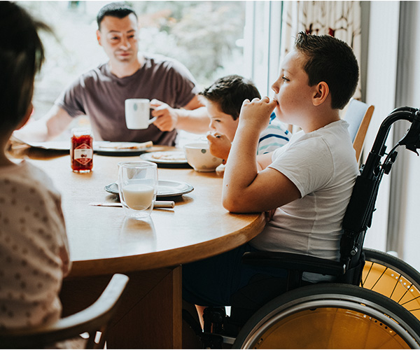 A boy sitting in a wheelchair at a table, eating a meal with his family. 