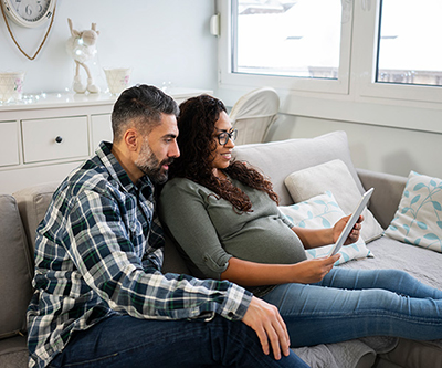 A man and a pregnant woman sitting together on a couch, looking at something on the tablet the woman is holding.