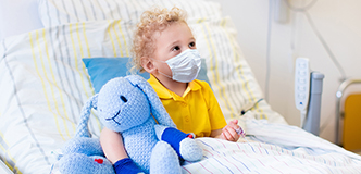 Masked child sitting in hospital bed holding a stuffed toy.