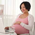 Pregnant woman, seated before laptop, with glass of water on desk.