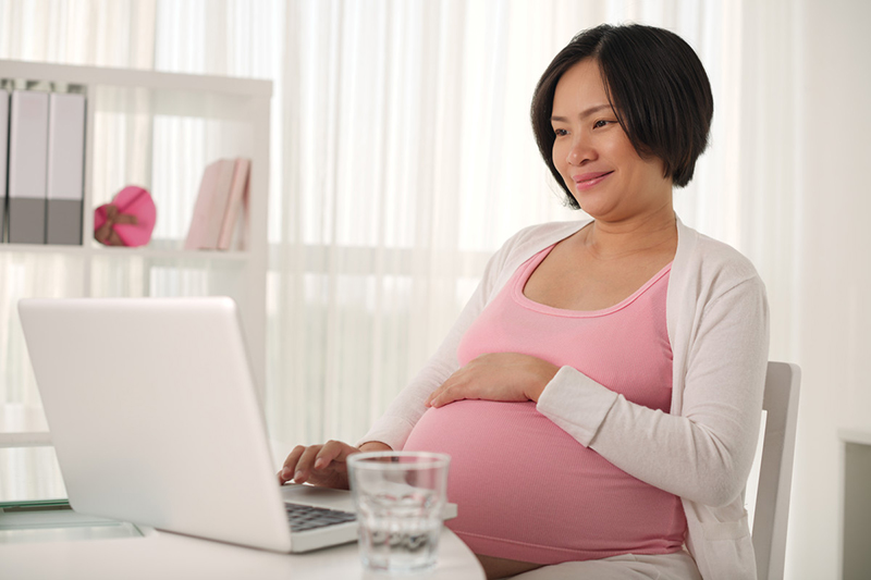 Pregnant woman, seated before laptop, with glass of water on desk.