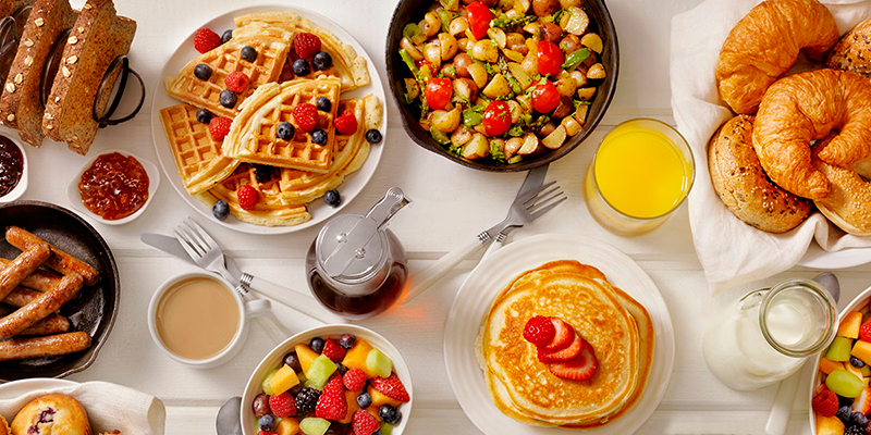 A white table with an assortment of beverages and plates of breakfast food, including waffles, pancakes, fruit, and croissants.