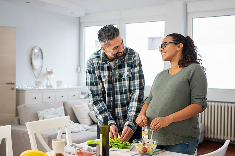 Man and pregnant woman standing over table preparing salad.