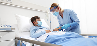 Masked child lying in hospital bed while healthcare provider checks oxygen level on a finger pulse oximeter.