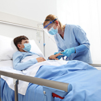 Masked child lying in hospital bed while healthcare provider checks oxygen level on a finger pulse oximeter.