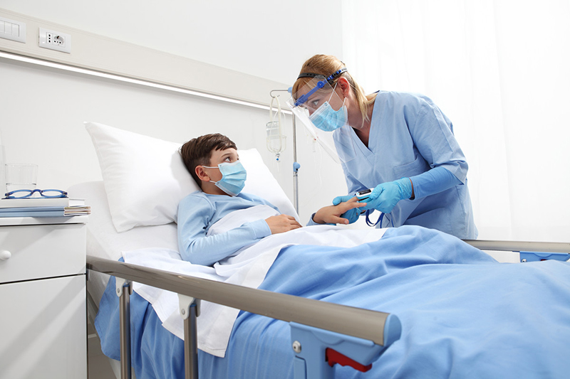 Masked child lying in hospital bed while healthcare provider checks oxygen level on a finger pulse oximeter.