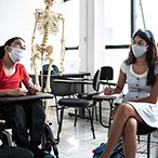 Children are seated in a classroom, including a boy in a wheelchair and a girl at a desk. Both are wearing masks.