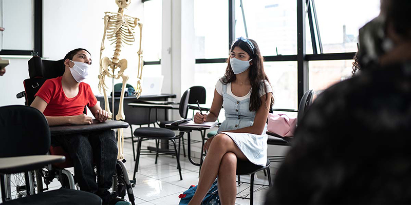 Children are seated in a classroom, including a boy in a wheelchair and a girl at a desk. Both are wearing masks.