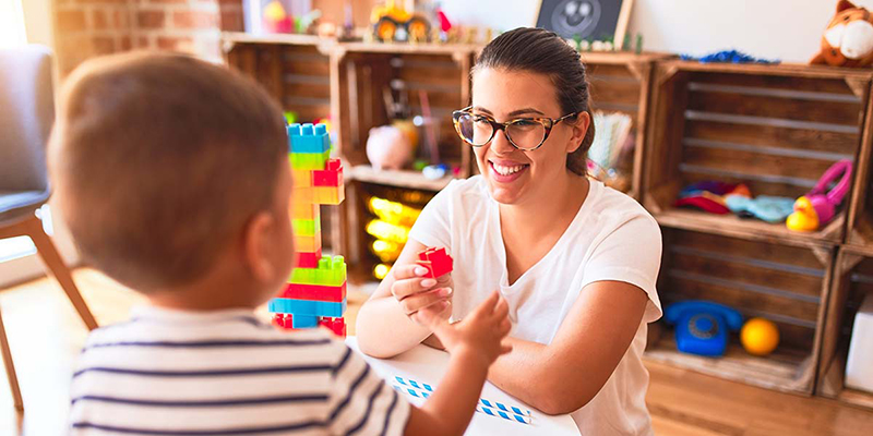 A child and an adult are inside a home environment playing with toy blocks across a table.