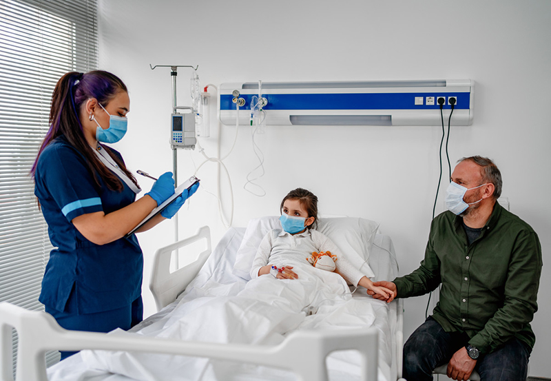 A girl is in a hospital bed flanked by a healthcare provider, who is filling out a chart, and her father, who is sitting down and holding her hand.