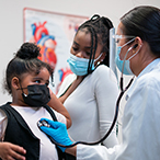 A healthcare provider examines a young girl with a stethoscope as her mother looks on. Everyone is masked.