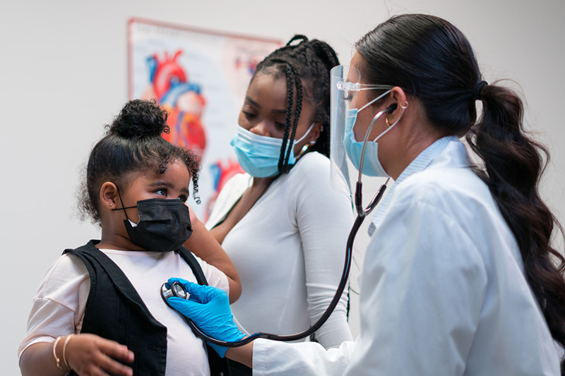 A healthcare provider examines a young girl with a stethoscope as her mother looks on. Everyone is masked.