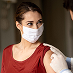 A woman receives a vaccination in her upper arm. She and the person administering the shot are masked.