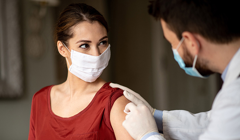 A woman receives a vaccination in her upper arm. She and the person administering the shot are masked.