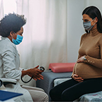 A pregnant woman holds her belly as she sits across from her healthcare provider, who is holding a stethoscope. Both are wearing masks.