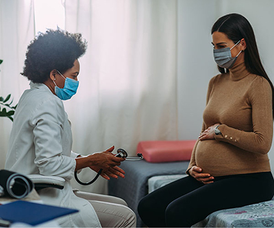 A pregnant woman holds her belly as she sits across from her healthcare provider, who is holding a stethoscope. Both are wearing masks.