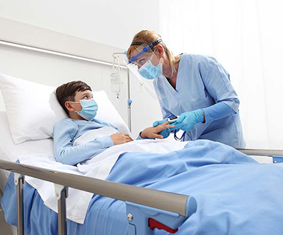 A masked boy in a hospital bed looks at his healthcare provider, who is checking his vital signs. She is wearing a mask and face shield.