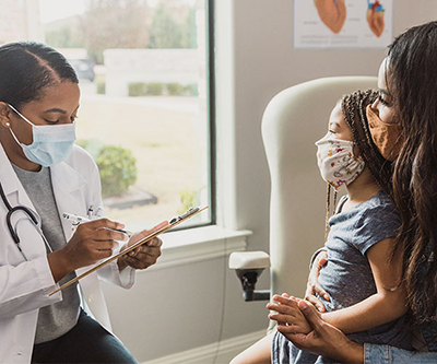 A healthcare provider takes notes across from her patient—a young girl and her mother. Everyone is wearing masks.