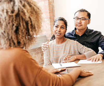 A man wraps his arm around his partner as both meet with a counselor or healthcare provider, who has her back to the camera.