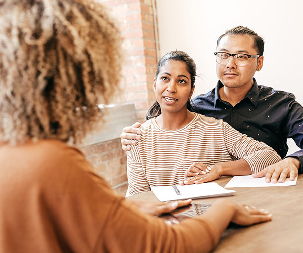 A man wraps his arm around his partner as both meet with a counselor or healthcare provider, who has her back to the camera. 