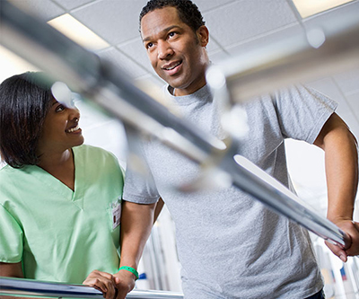 A therapist helps a man as he smiles during a therapy session. He is gripping two parallel bars.