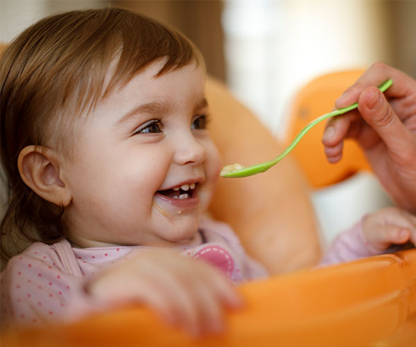 An infant smiles in a high chair as an adult, who is not visible, feeds her with a green spoon.