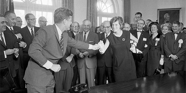 A black and white photo of President John F. Kennedy reaching for a pen in a crowded room.