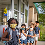 A group of elementary school-aged children are lined up outside a building. They are all wearing cloth face masks.