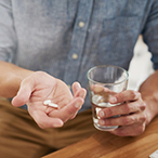 A man seated at a table holds pills in one open hand while holding a glass of water in the other hand.