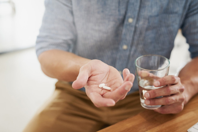 A man seated at a table holds pills in one open hand while holding a glass of water in the other hand.