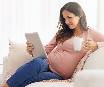 A smiling pregnant person reclines on a sofa, holding a white mug and a computer tablet.