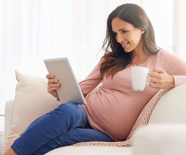 A smiling pregnant person reclines on a sofa, holding a white mug and a computer tablet. 