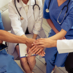 Medical staff wearing white coats and scrubs sit in a semicircle with their hands stacked in the middle.