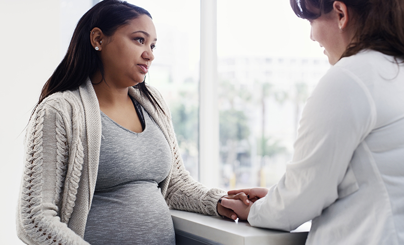A healthcare provider wearing a white coat holds the hand of a pregnant person while talking with her.
