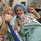 A blonde woman wearing a mask crouches next to a smiling girl lying on a hospital bed.