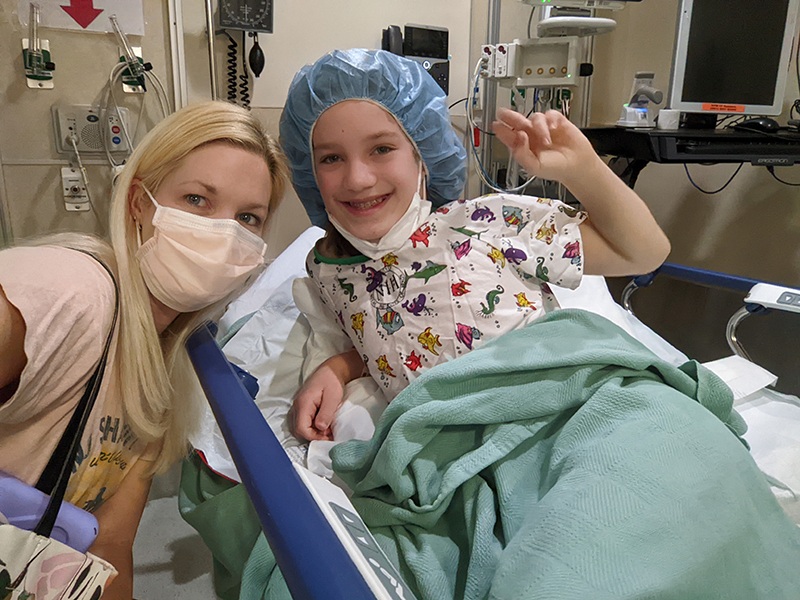 A blonde woman wearing a mask crouches next to a smiling girl lying on a hospital bed.