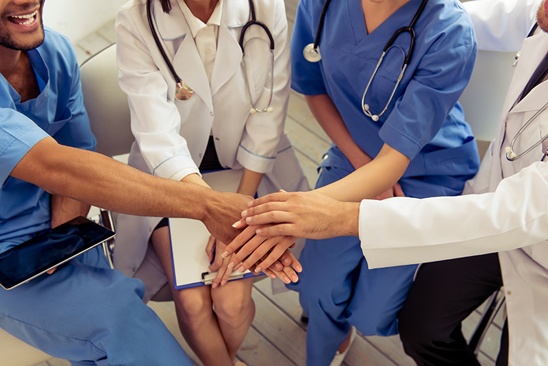 Medical staff wearing white coats and scrubs sit in a semicircle with their hands stacked in the middle.