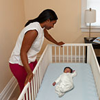 In the parent’s bedroom, a mother smiles over her sleeping baby, who is asleep on the back in a crib. The baby’s separate sleep area adheres to safe sleep guidelines.