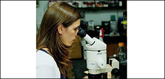 A woman with brown hair wearing a white jacket and white lab gloves peers into a microscope. Bottles and other scientific research equipment are blurred in the background.