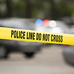 Yellow tape with black text stating “Police line do not cross” in front of out-of-focus police cars and greenery.